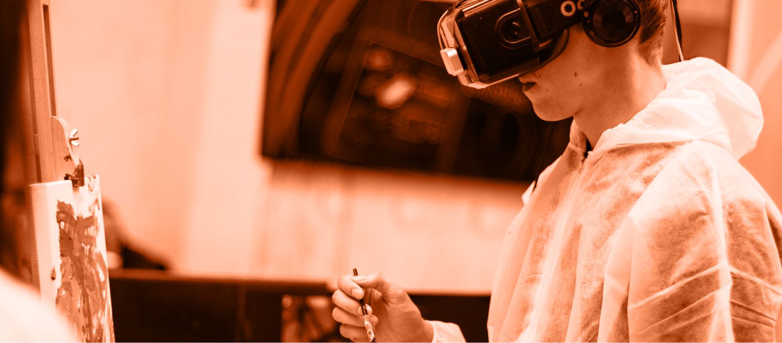 A boy wearing virtual reality goggles and plastic overalls, about to paint on a canvas.